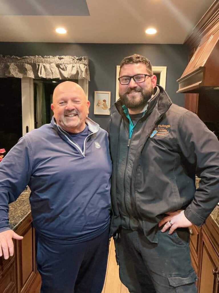 Two men stand side by side in a kitchen, smiling at the camera. One wears a blue pullover, the other a black jacket with an embroidered logo.