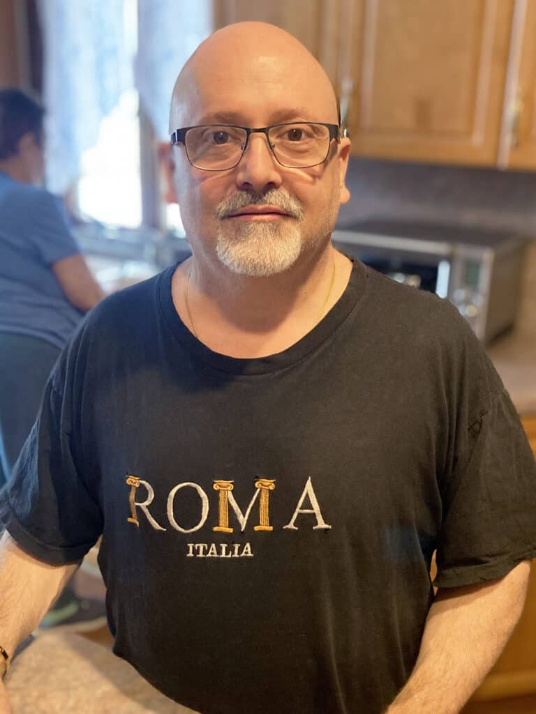 A man with glasses and a goatee stands in a kitchen wearing a black "ROMA ITALIA" t-shirt; another person is visible in the background.
