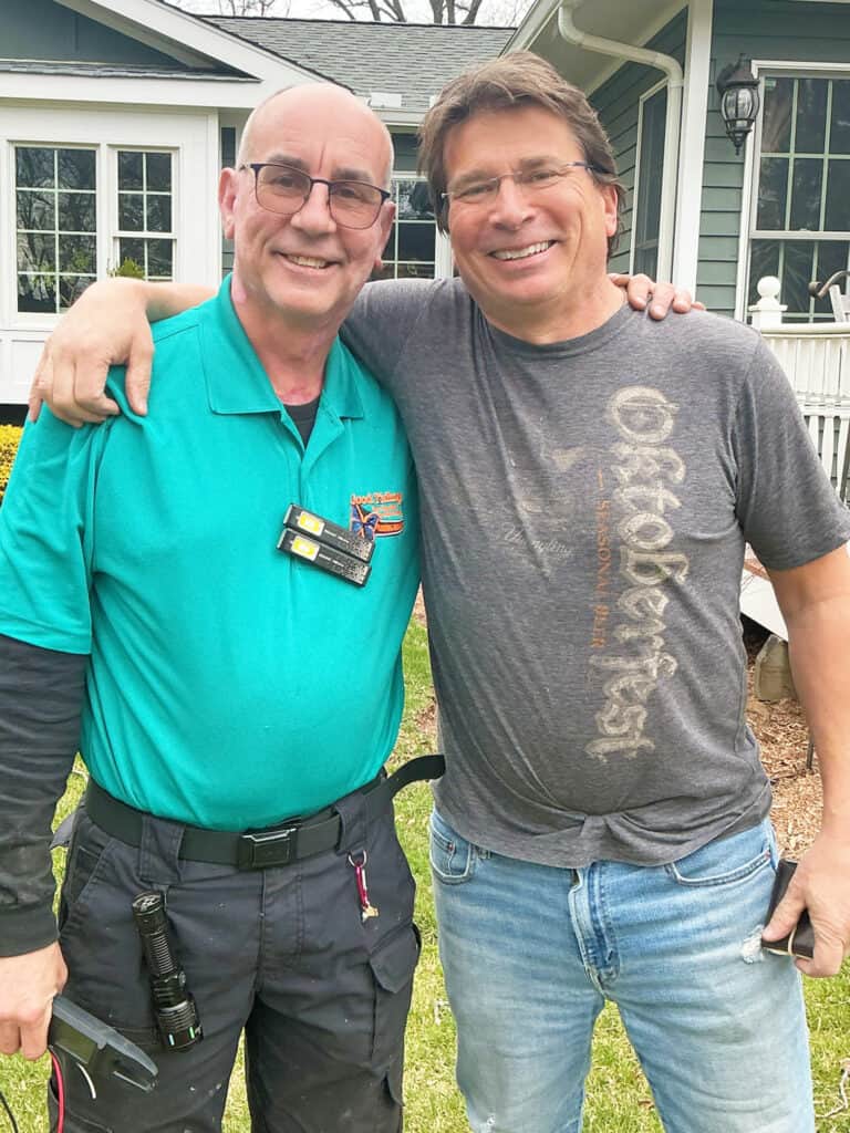 Two men stand outside a house, smiling with their arms around each other. One wears a teal polo and work pants; the other wears a gray T-shirt and jeans.