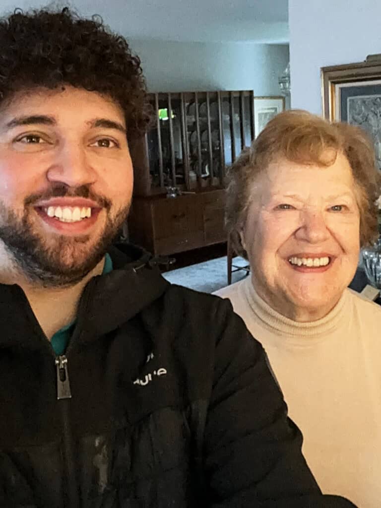 A young man and an older woman are standing indoors and smiling at the camera.