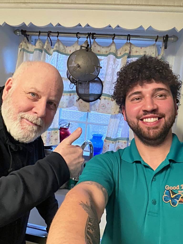 Two men pose for a selfie in a kitchen; one gives a thumbs up while the other smiles at the camera. A window with curtains and dishes are visible in the background.