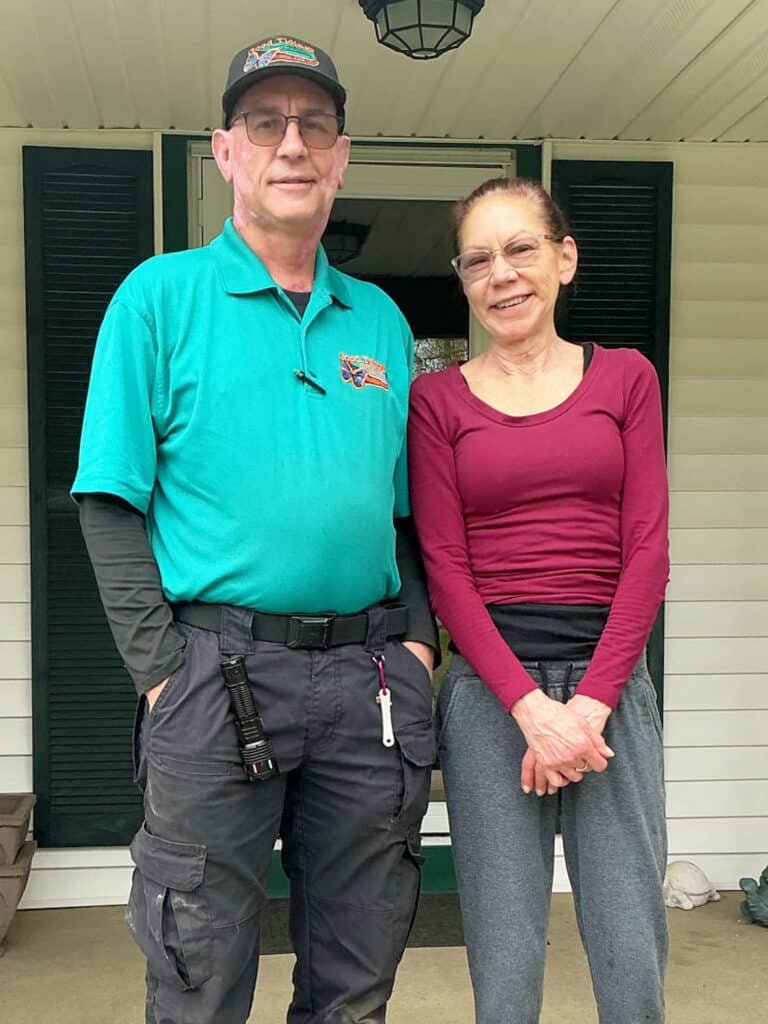 Two adults stand side by side on a porch in front of a doorway. The man wears a teal shirt and hat; the woman wears glasses, a maroon top, and gray pants.