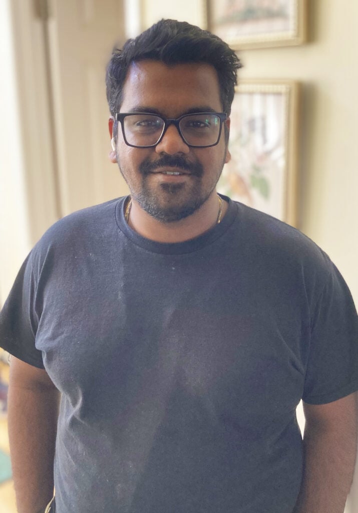 A man with dark hair, beard, and glasses wearing a black t-shirt stands indoors, looking at the camera and smiling slightly.