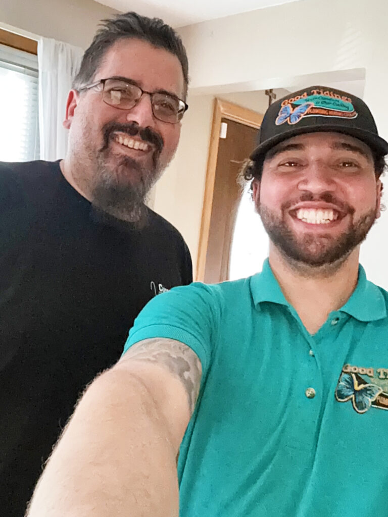 Two men smiling indoors; one wears a black shirt and glasses, the other wears a teal polo and matching cap with embroidered logos.