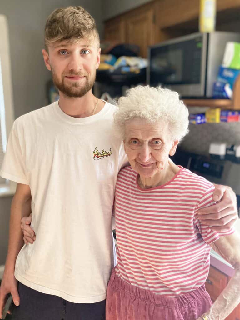 A young man and an elderly woman stand together in a kitchen, both smiling at the camera. The woman wears a striped shirt and the man wears a white t-shirt.