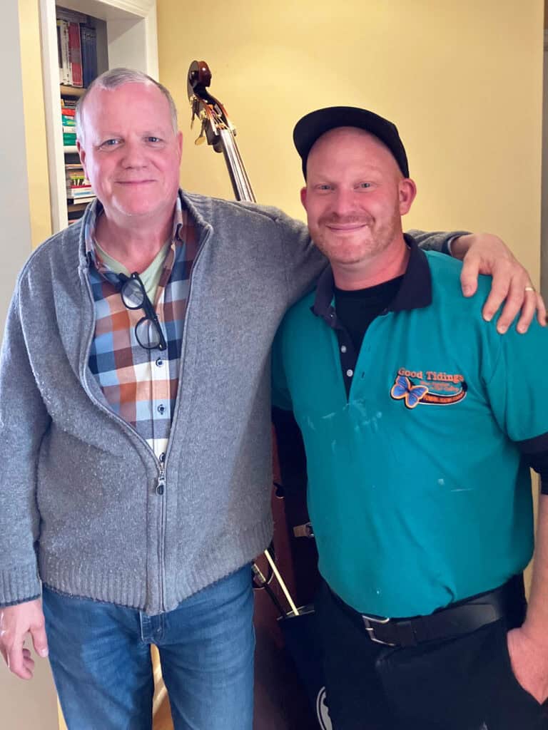 Two men stand side by side indoors, one wearing a grey sweater and glasses, the other in a teal polo and black cap. A string bass is visible in the background.