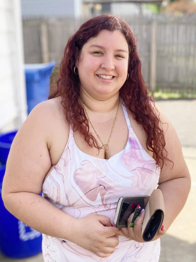A woman with long curly hair is standing outside, smiling, and holding a phone and a beige clutch. She is wearing a sleeveless white and pink dress.