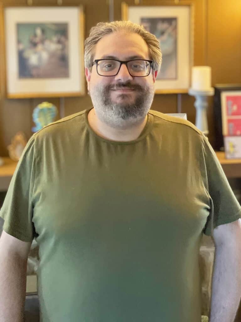 A man with glasses and a beard wearing an olive green T-shirt stands indoors in front of a blurred background with framed art and decorative items.