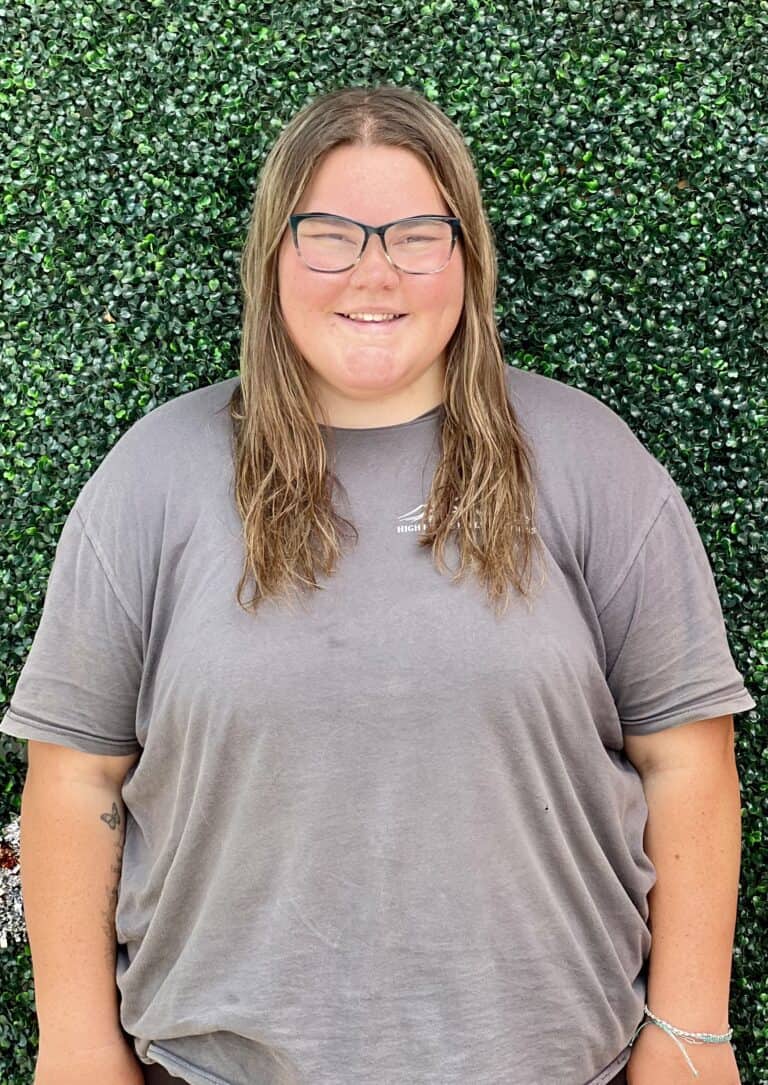 Meet Our Team 6 A young woman with glasses smiling in front of a lush green hedge, promoting High Plains K-9 Connections, emphasizing training, therapy dog programs, and community service with dogs.