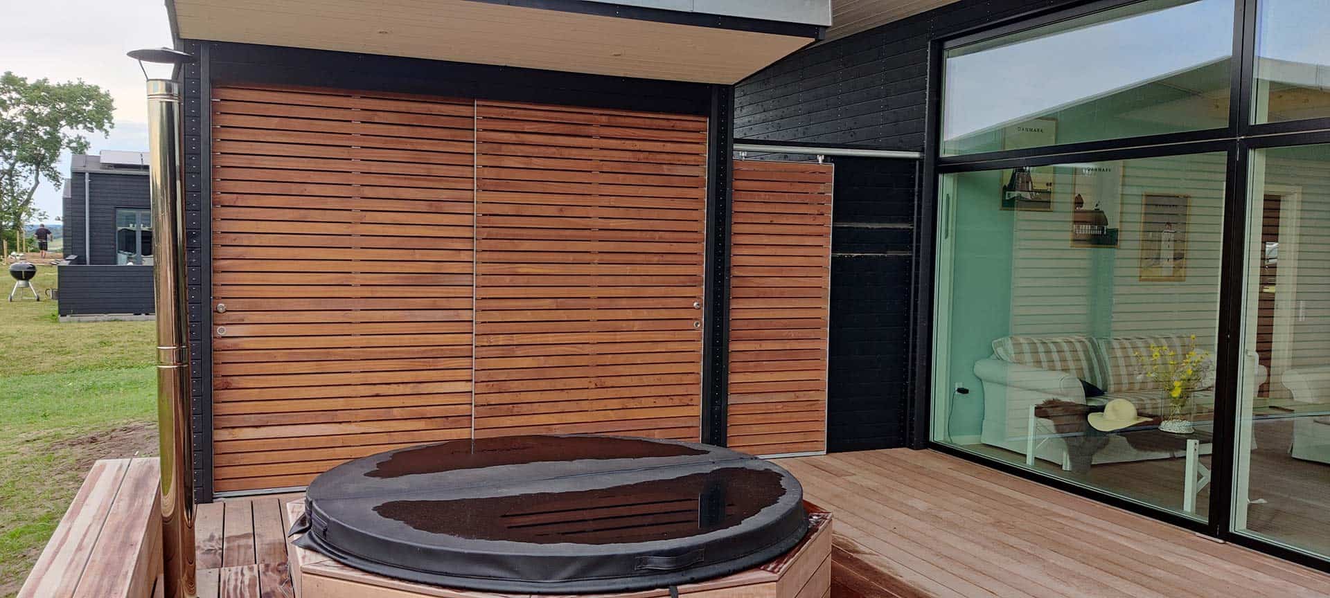 Udendørs facade med trælameller og en indbygget detalje i bunden. Foran ses en spa og træterrase.