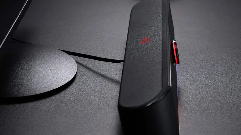 Side profile of the black Redragon GS560 soundbar on a dark desk next to a monitor stand