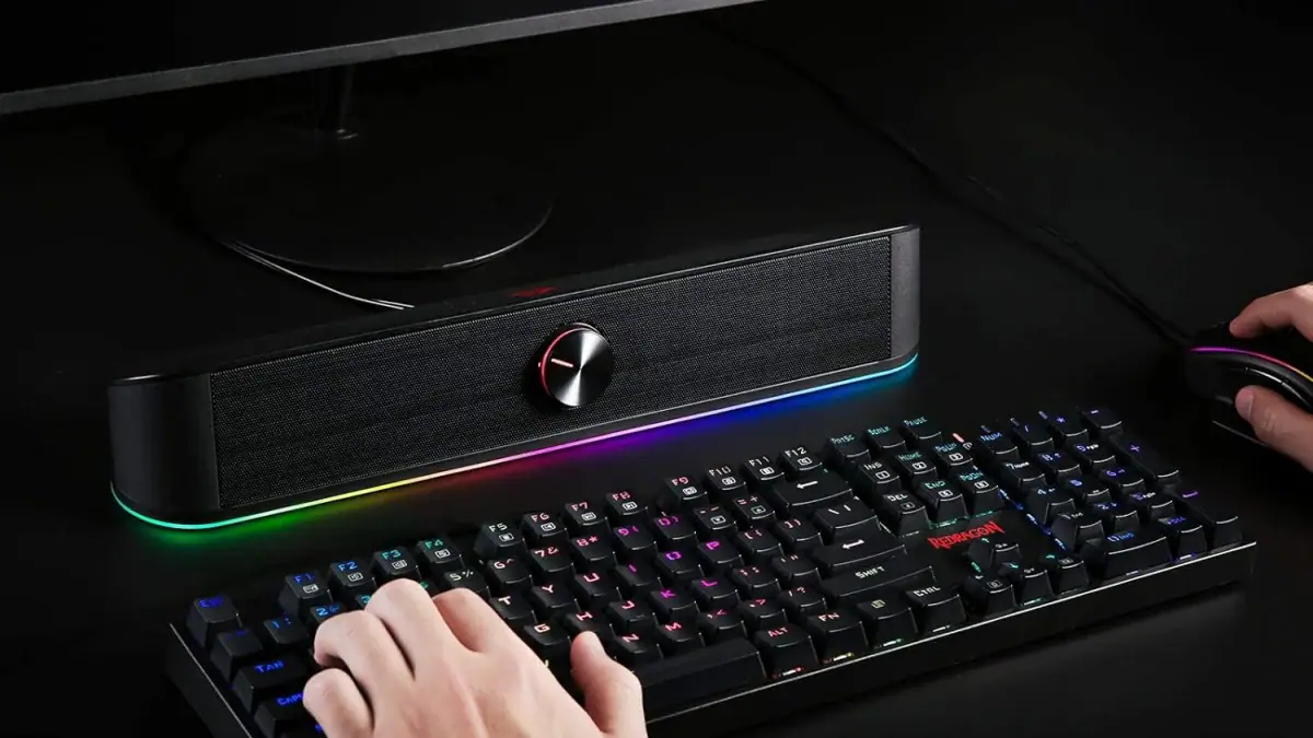 Redragon GS560 soundbar with its RGB light bar glowing under a monitor, behind a mechanical gaming keyboard