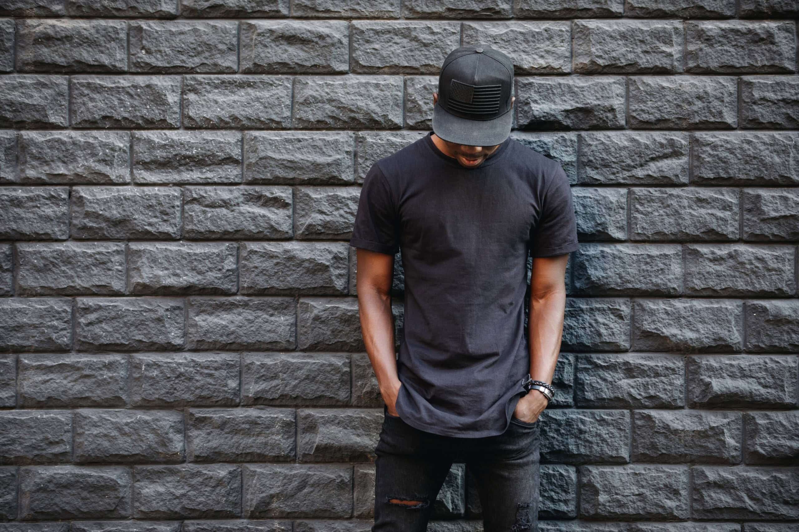 Young man wearing black T-shirt and cap standing against gray brick wall, casual street style.