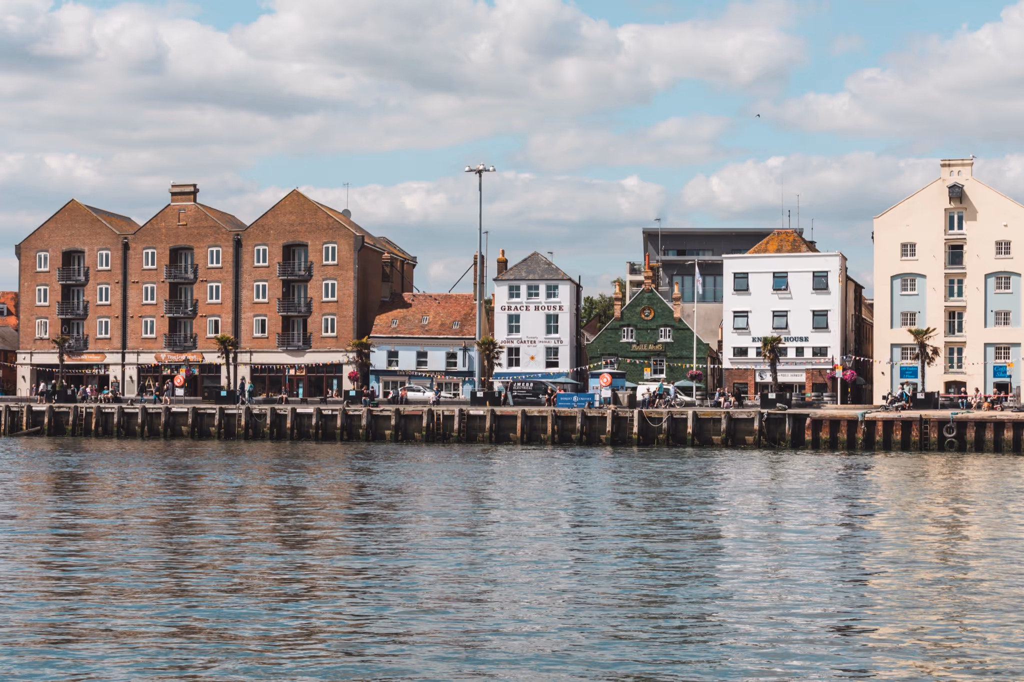 Waterfront view of several brick and white buildings with shops and a hotel, lined along a pier, reflected in calm water under a partly cloudy sky.