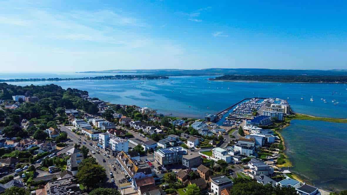 Aerial view of lilliput in Dorset coastal town with houses, roads, a marina, and blue water under a clear sky. Boats are moored in the harbour, and greenery surrounds the residential area.