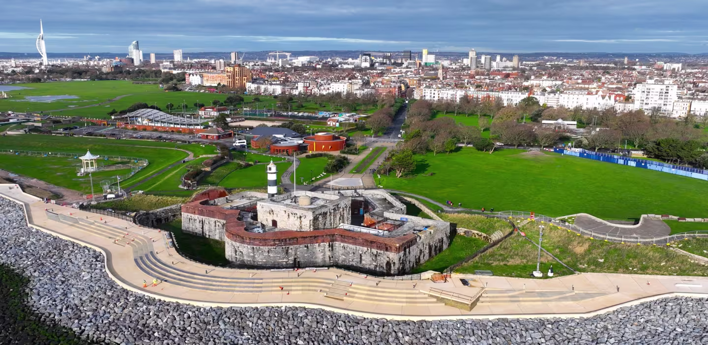 Aerial view of a historic stone fort with a lighthouse by the sea, set beside a large green park and a cityscape in the background under a partly cloudy sky.