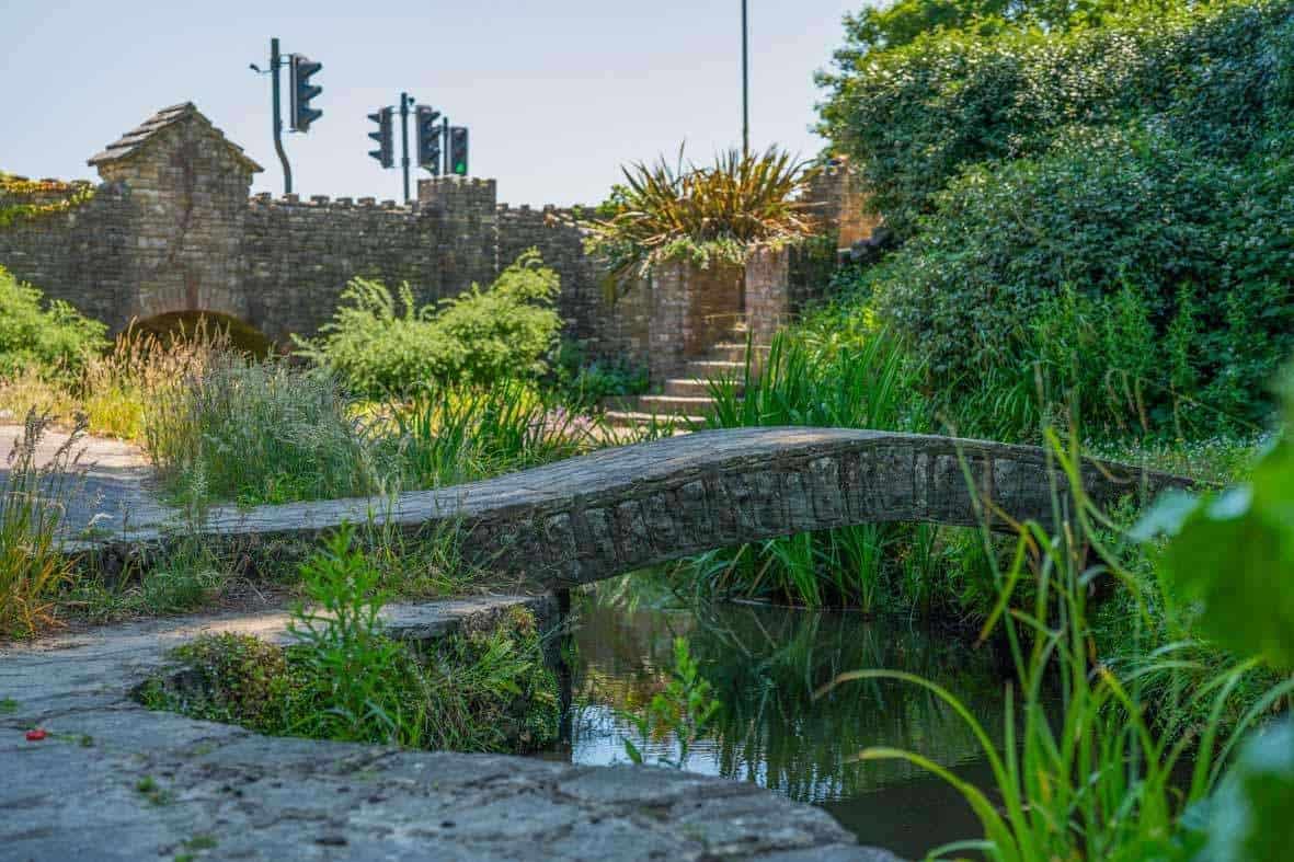 A small stone footbridge crosses a narrow stream in a lush garden, with a stone wall and traffic lights visible in the background.