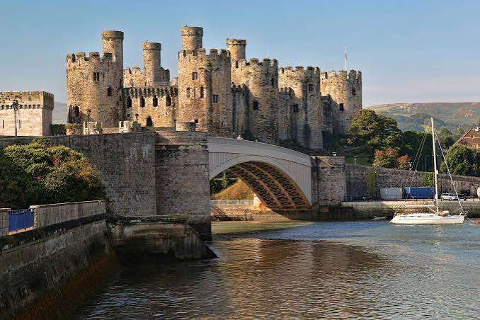 A stone castle with multiple round towers stands behind a large arched bridge over a river. A sailing boat is moored nearby, and trees and hills are visible in the background under a clear sky.
