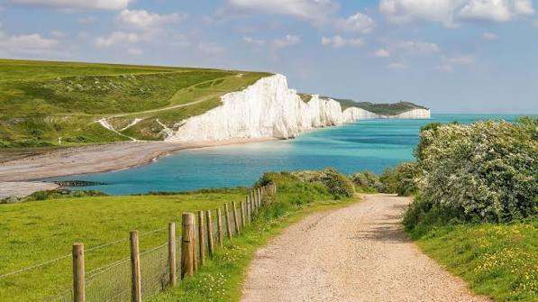 A dirt path leads through grassy fields towards white chalk cliffs by a turquoise sea, under a partly cloudy sky. DOver in ENgland