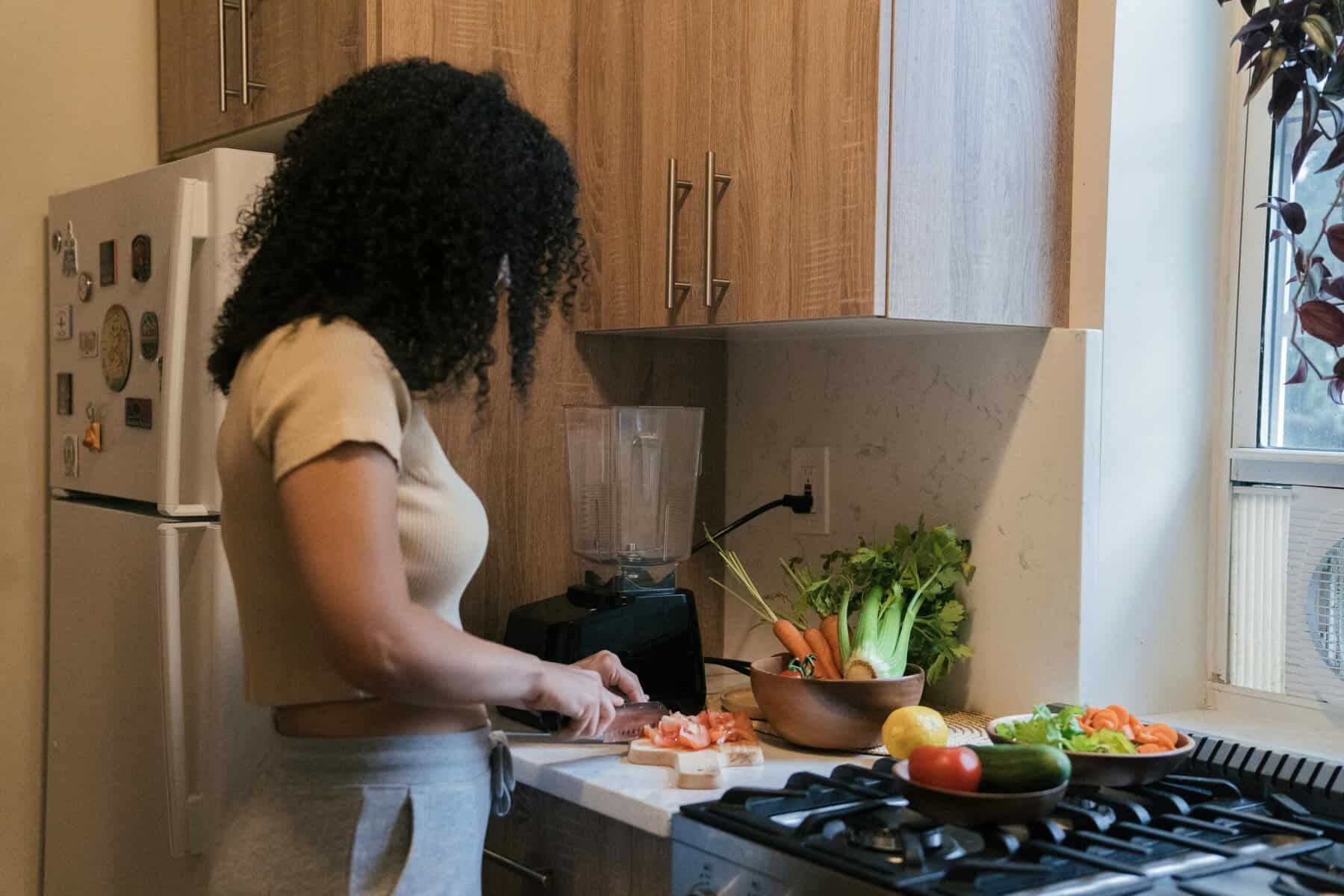 Woman with curly hair chopping vegetables on a kitchen counter surrounded by fresh carrots, celery, and tomatoes, preparing an anti-inflammatory meal