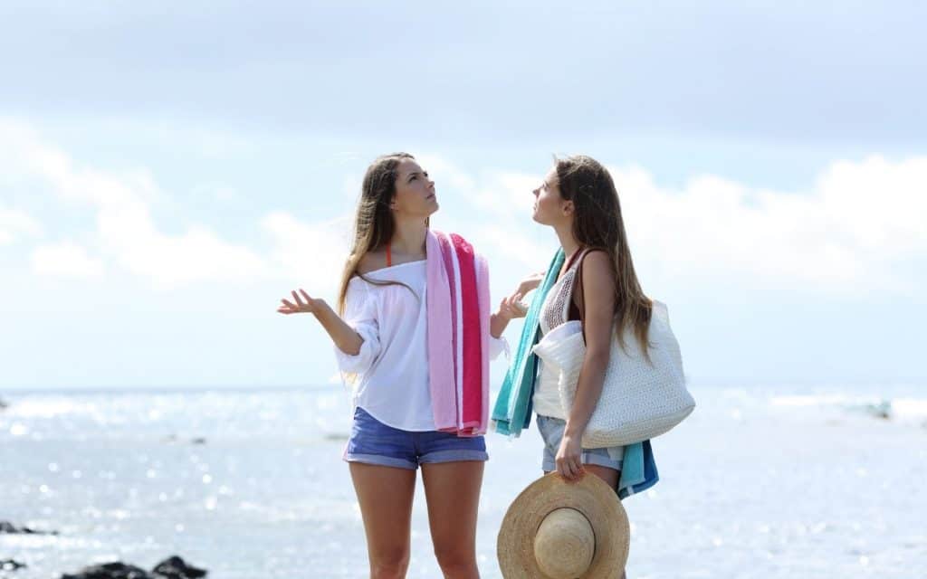 Sunny beach scene with two women having a conversation, holding towels and a hat.