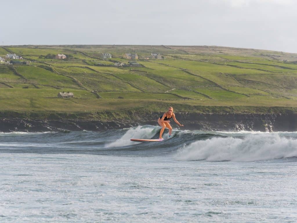 Surfer riding wave along rugged Irish coast with green fields and small houses in background.