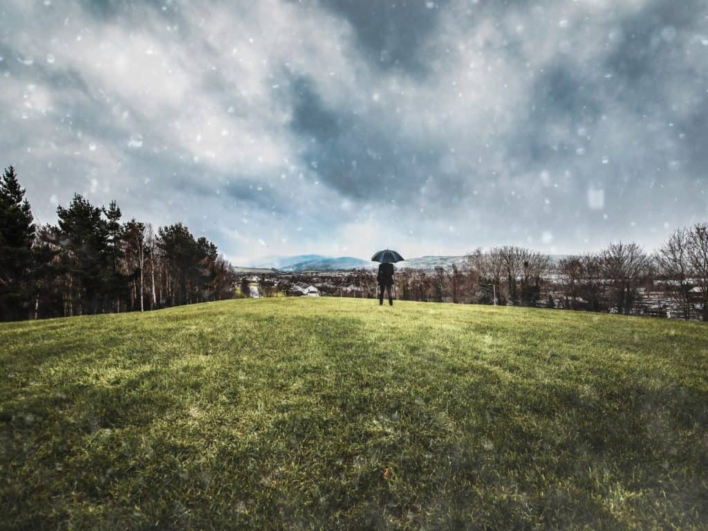 Grey sky with rain, person holding umbrella on lush green field in Ireland, scenic landscape.