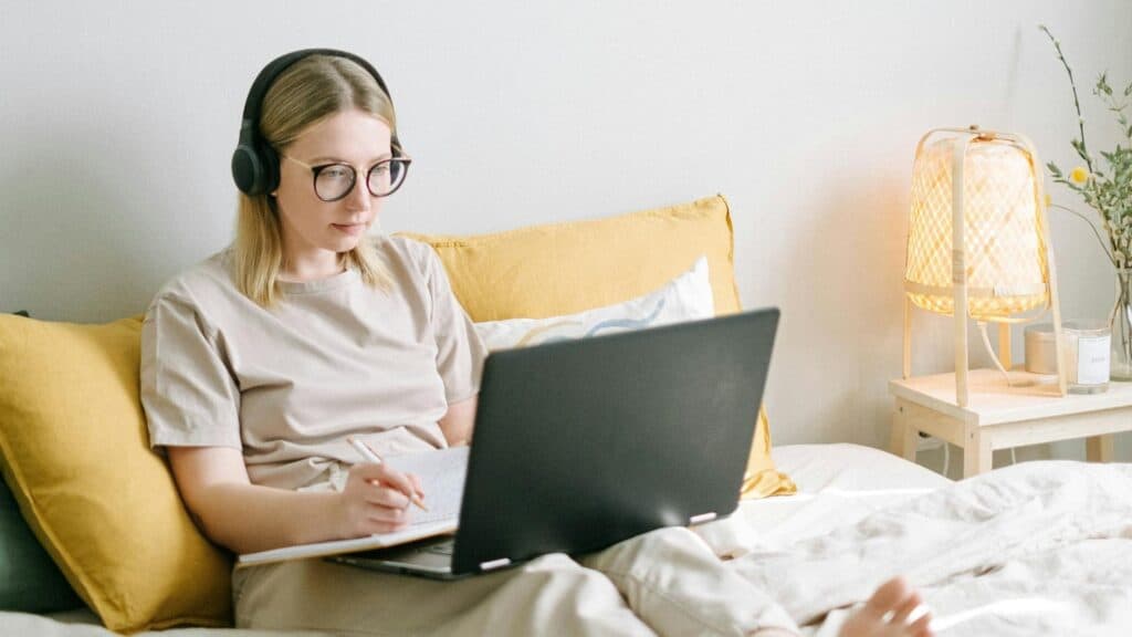 Young woman with headphones working on a laptop in bed, taking notes with a pen.