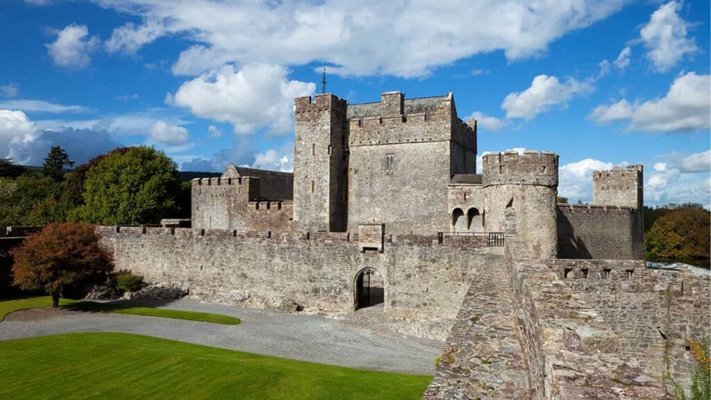 Medieval Irish castle surrounded by lush green trees under a bright blue sky with fluffy clouds.