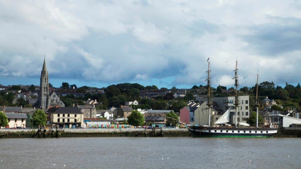 Sailing ship docked in a scenic Irish harbor town with colorful buildings and a church spire in the background.