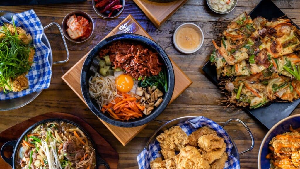 A variety of delicious Asian dishes served on a wooden table, including fried chicken, vegetable bibimbap, sushi, and stir-fried noodles.