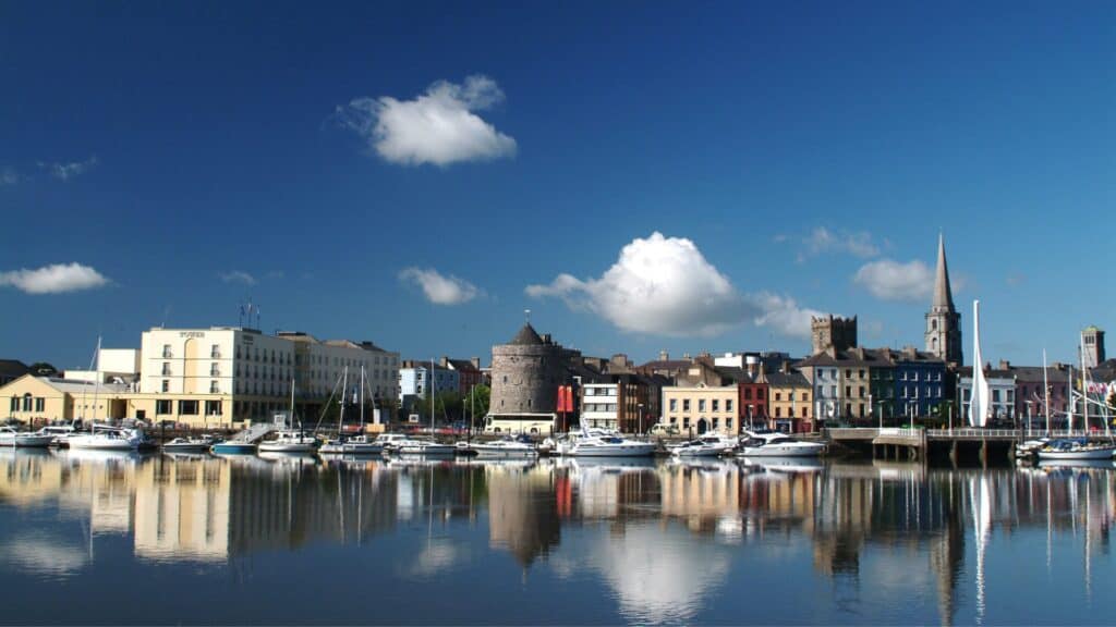 Colorful Irish harbor town with historic architecture, boats, and reflections on calm water under blue sky, showcasing Ireland's coastal beauty and culture.