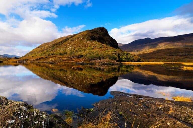 Serene Irish mountain reflected in tranquil lake with a blue sky and scattered clouds.