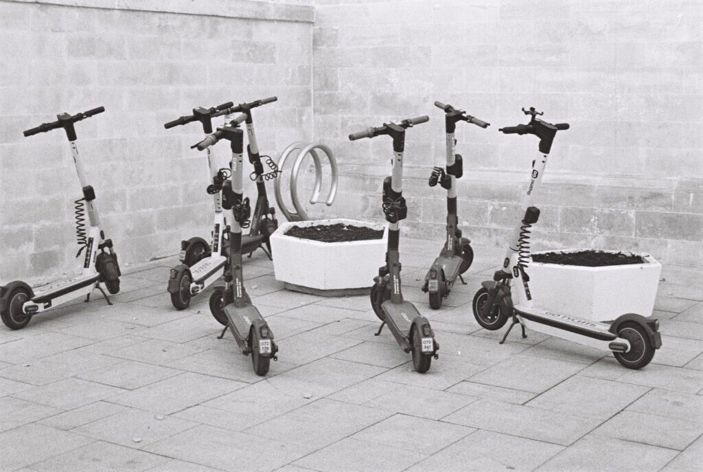 Black and white photo of several electric scooters parked beside concrete planters.