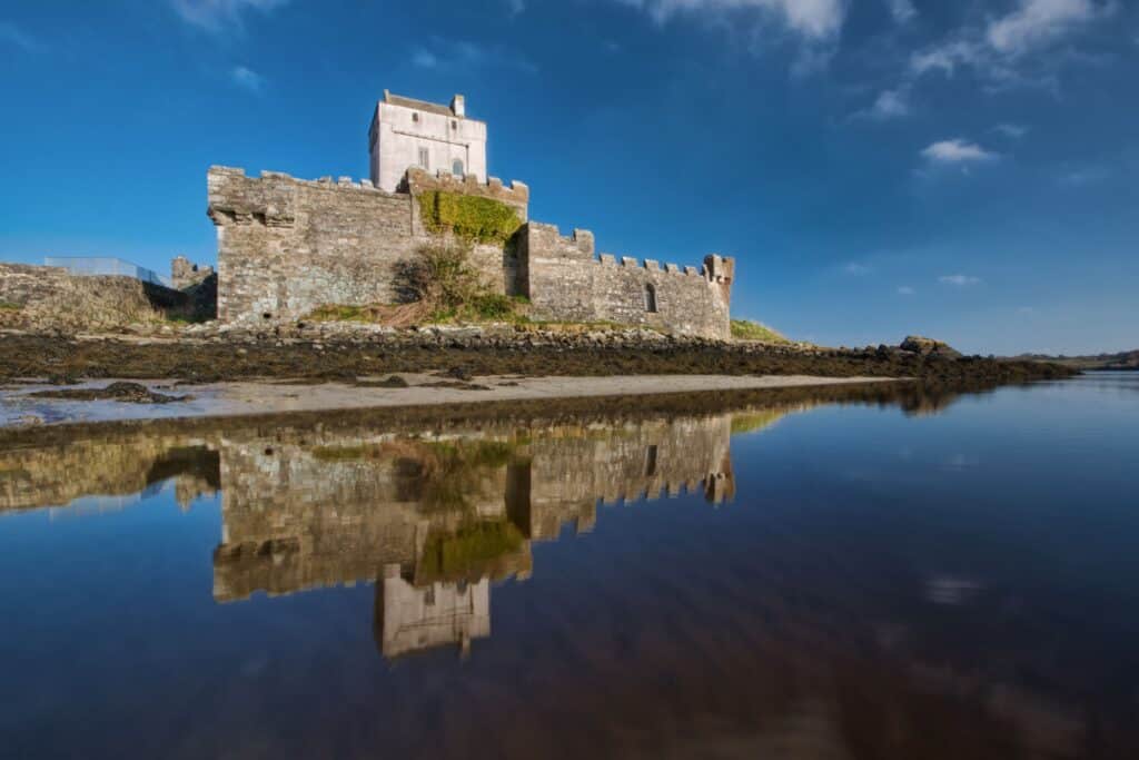 A stunning view of Doe Castle in County Donegal, Ireland reflecting in calm waters under a clear blue sky.