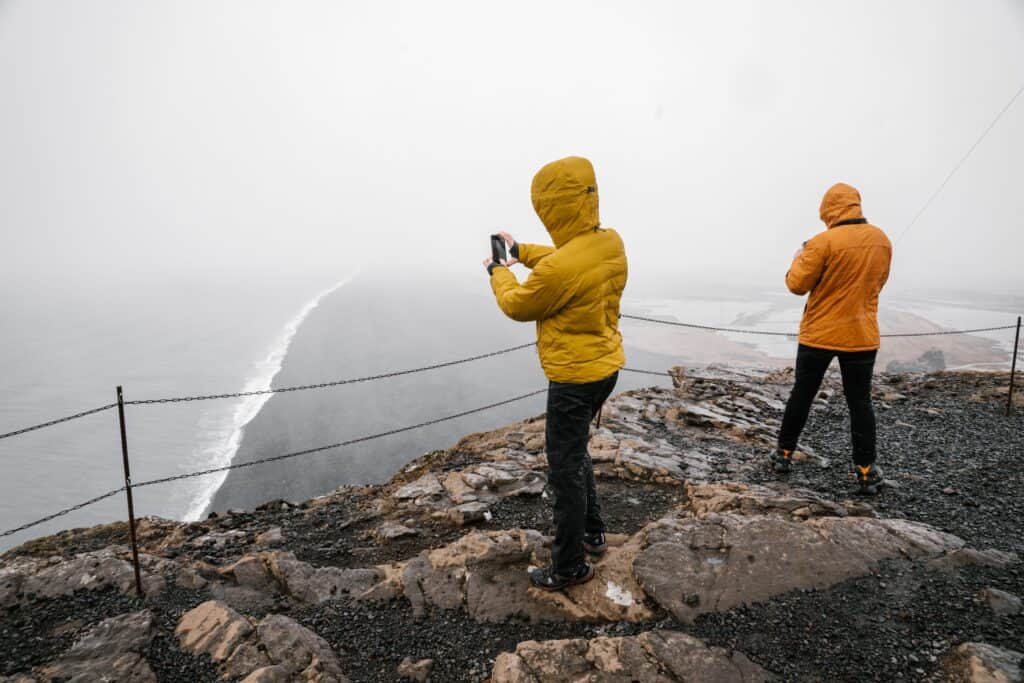 Two hikers in waterproof jackets capture photos on a foggy cliff overlooking the coastline.