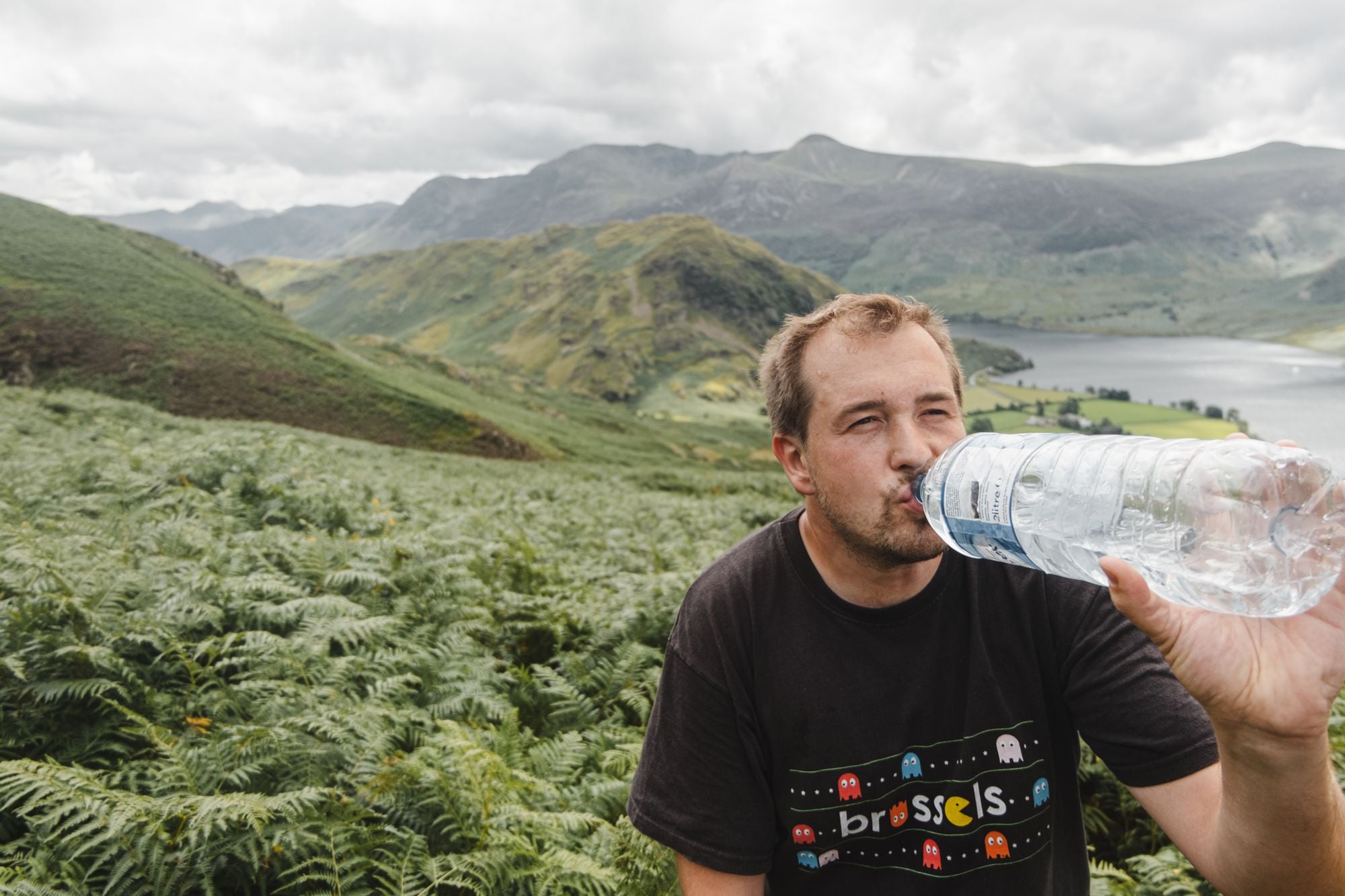 Adult man sipping water from bottle while resting near lush fern plants on slope of mountain on cloudy day in higlands