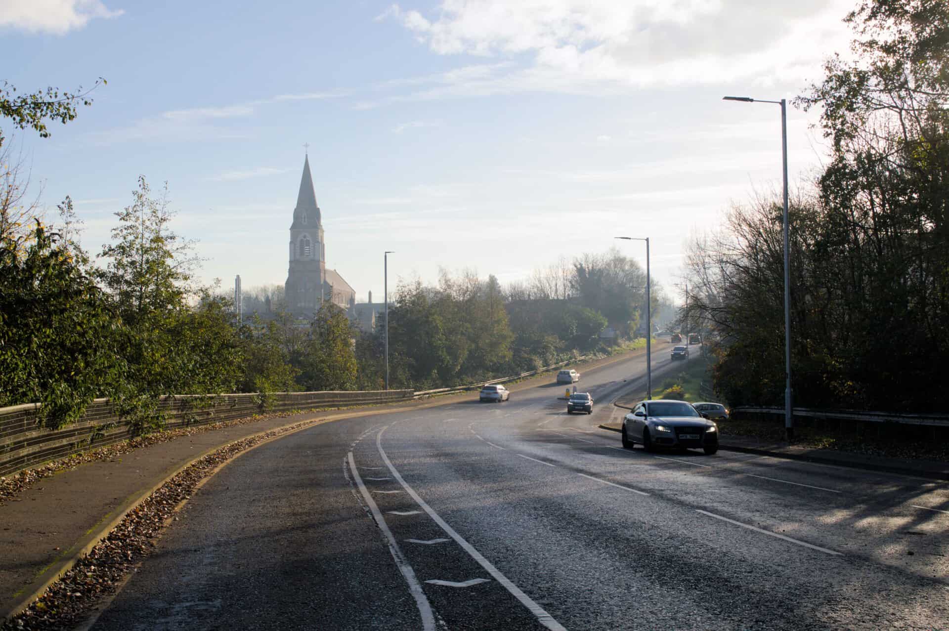 Irish road with church spire in the background and cars driving by.