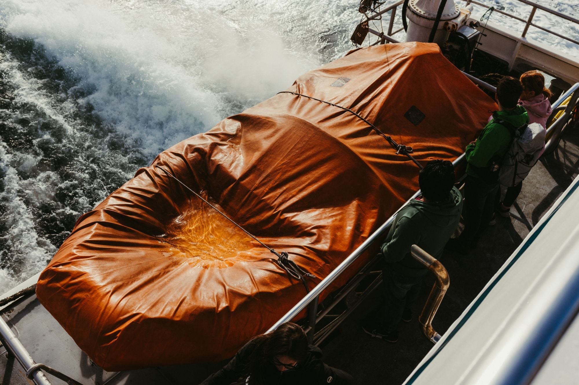 A group of people stands on a boat deck near an orange lifeboat off the coast of Ireland.