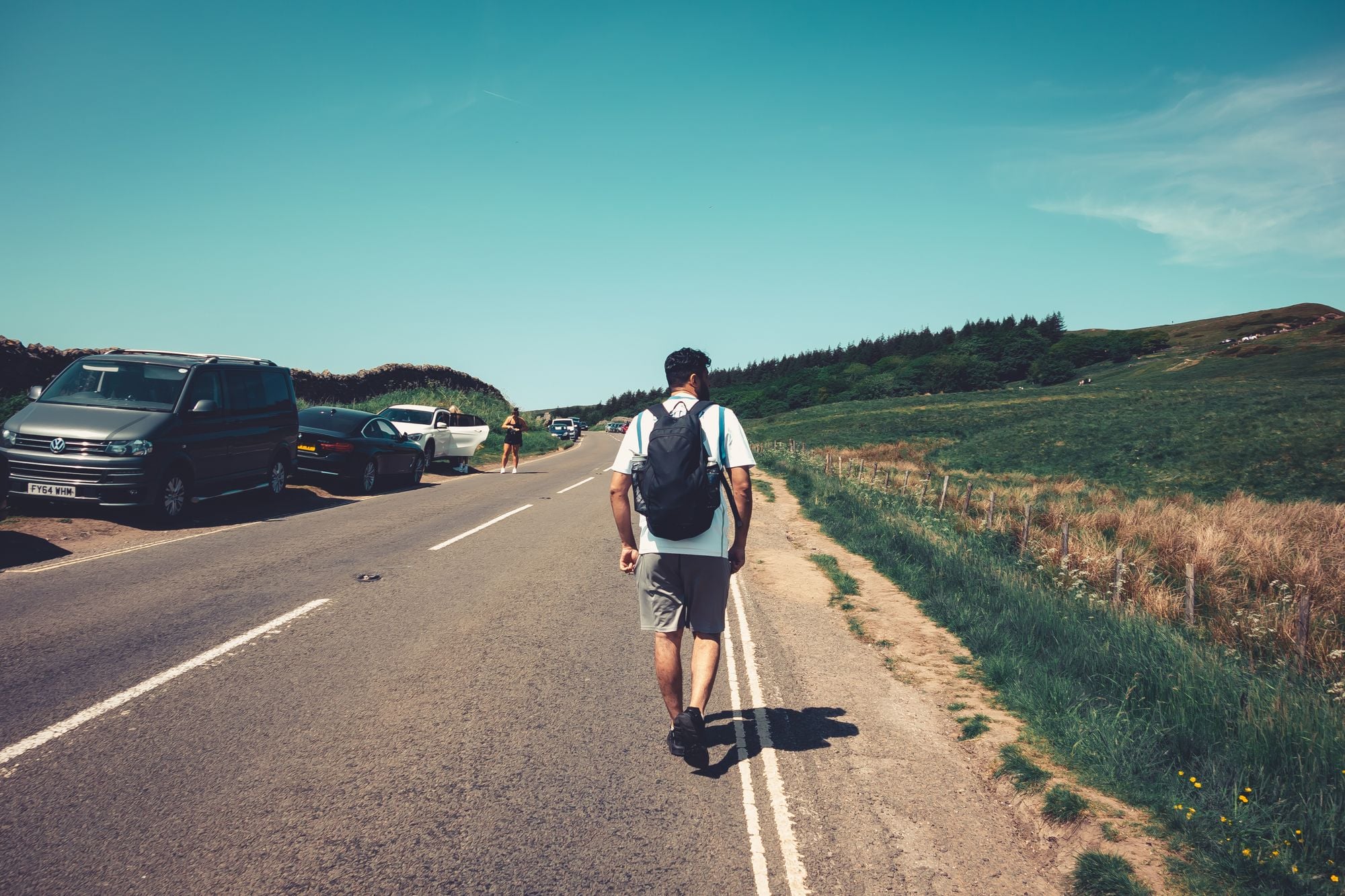 A man wearing a backpack walks along a scenic country road on a sunny summer day.