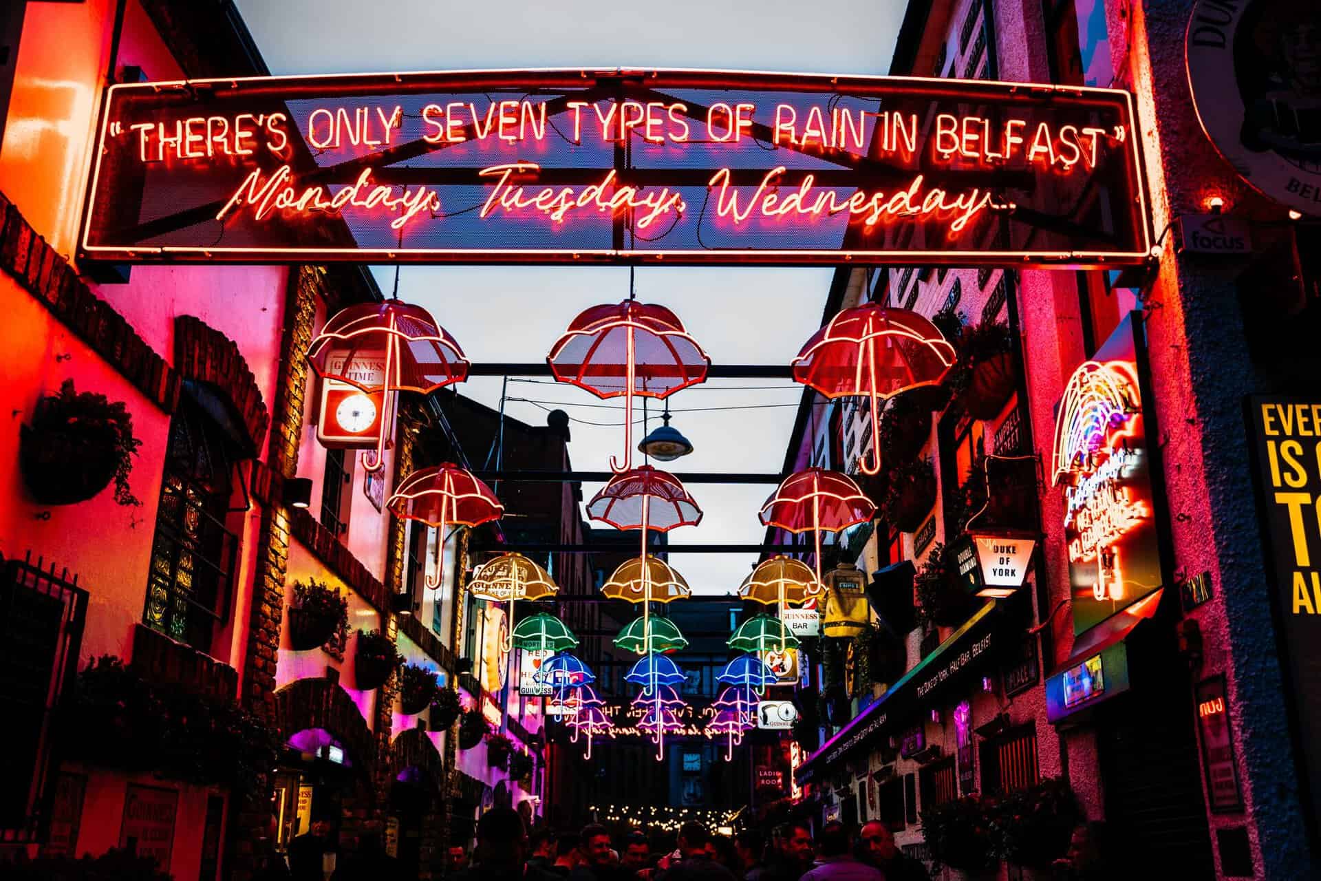 Bustling Belfast street illuminated by colorful neon umbrellas and a witty sign.