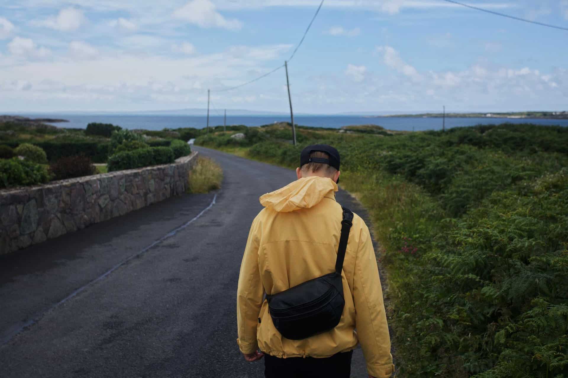 Man in yellow jacket walking on a scenic coastal road in Ireland, surrounded by lush greenery.
