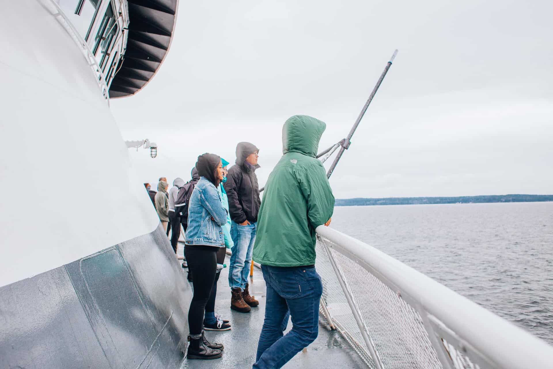 Passengers in jackets enjoy the view from a ferry deck on an overcast day, observing the calm waters.