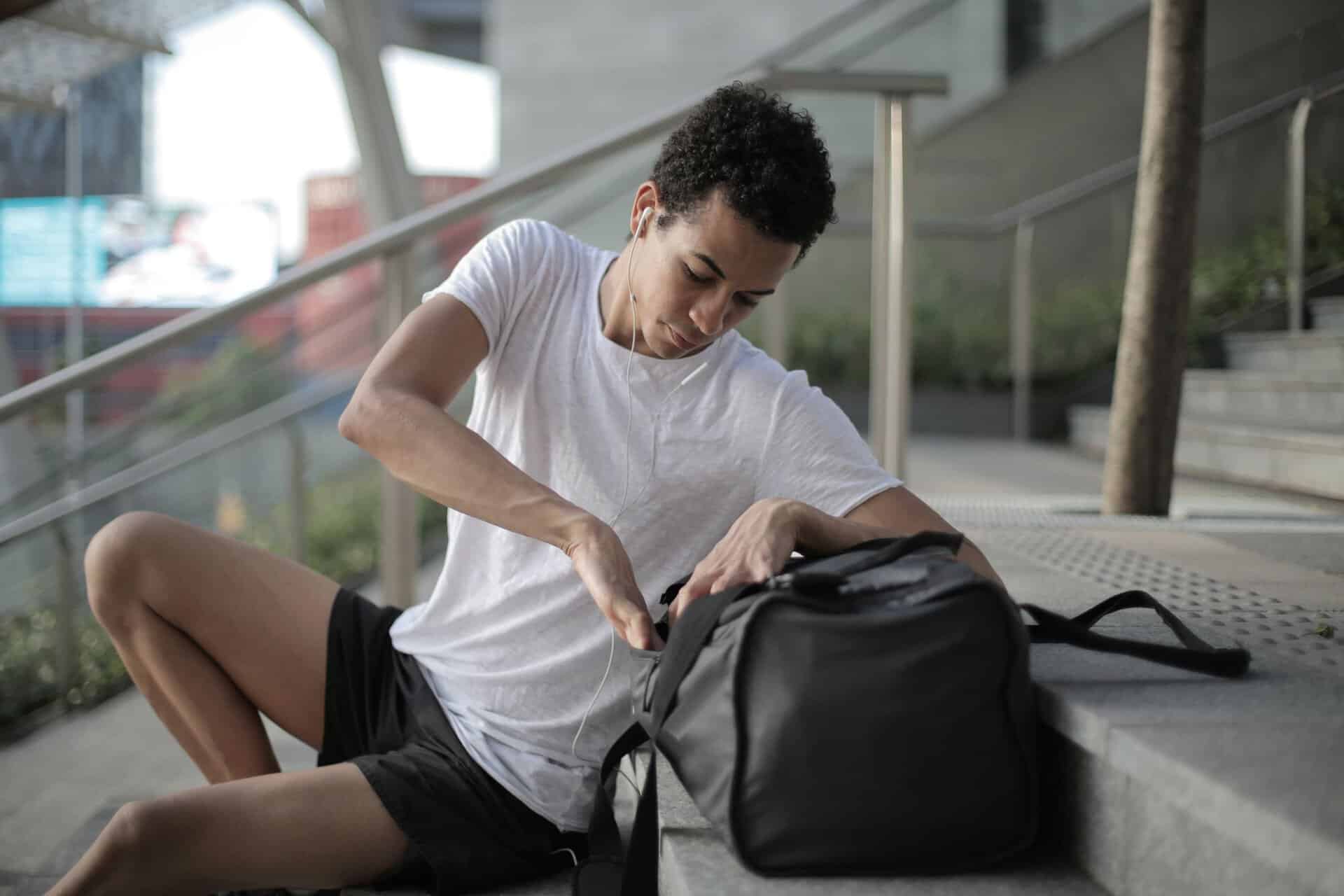 Serious thoughtful African American man in earphones and sports clothes searching in bag pocket device while sitting on stairs and listening to music at entrance of contemporary building in downtown
