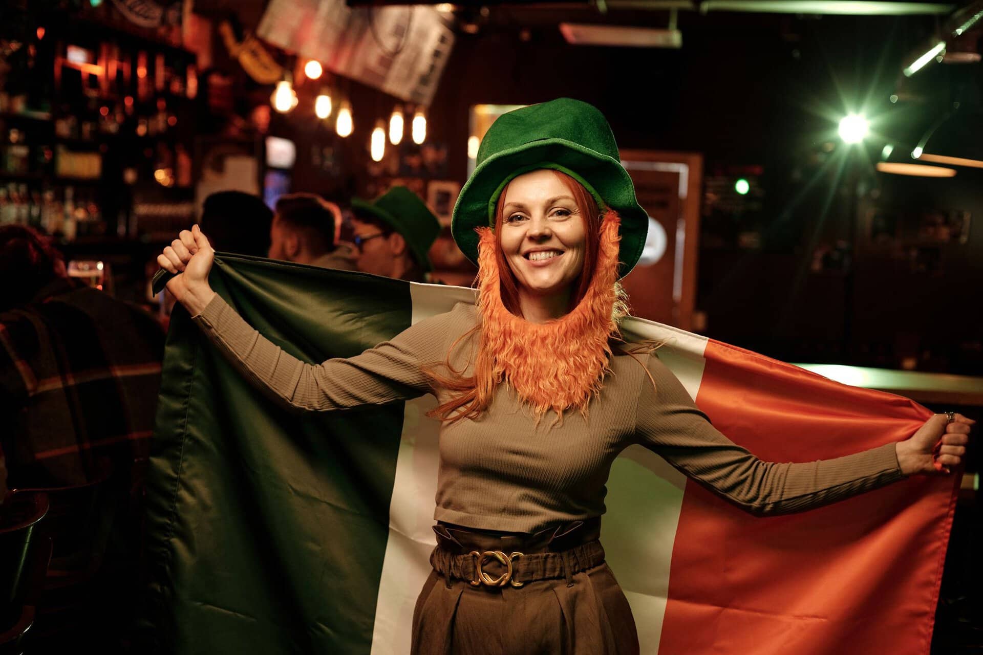 Smiling woman in green hat holding Irish flag at St. Patrick's Day celebration in a lively pub.