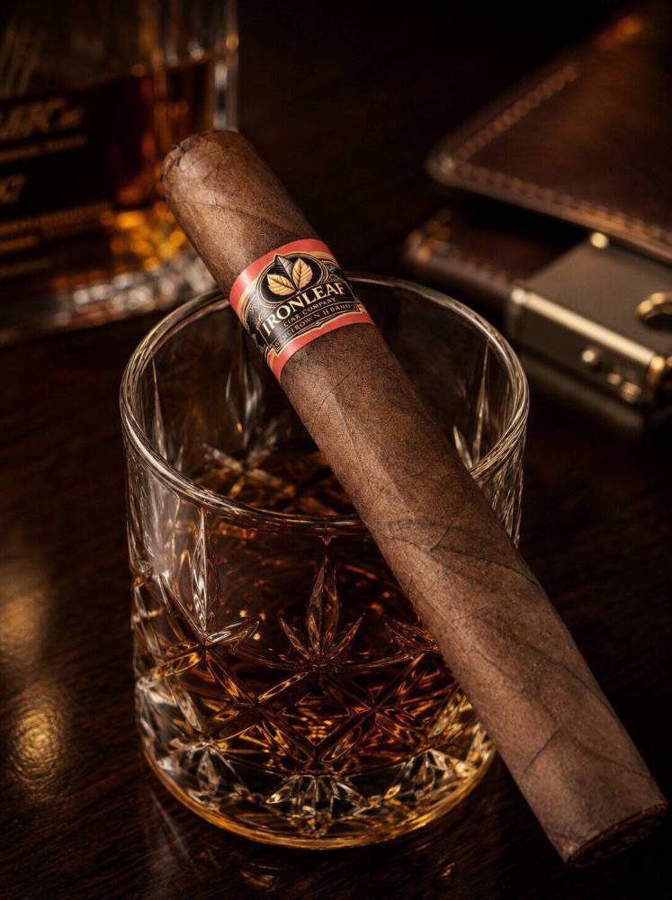 A premium Iron Leaf cigar rests across a crystal whiskey glass filled with amber liquor on a dark wooden table. The scene is warmly lit with a luxurious lounge atmosphere, featuring rich brown tones, soft reflections, and blurred background accessories that create an elegant, sophisticated cigar-and-whiskey presentation.