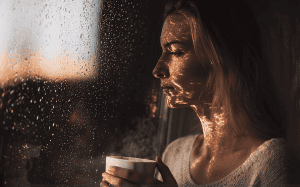 A woman looks out a rain-streaked window at dusk, holding a warm cup as soft light reflects across her face.