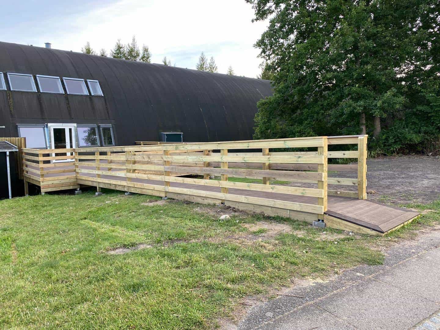 Træhegn og udendørstrappe foran en bygning med grønne omgivelser.