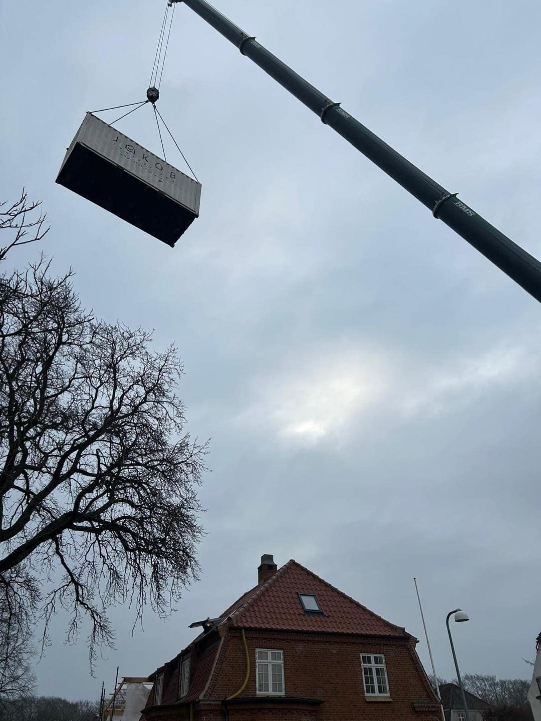 Kran løfter en stor byggecontainer over et hus under opførelse.