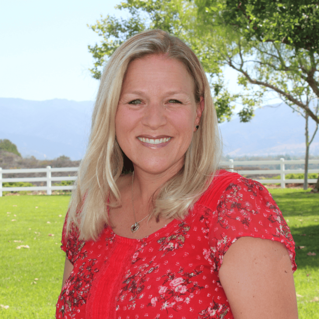 Jodi Snowdon smiling outdoors in a red floral top with trees and mountains behind her.